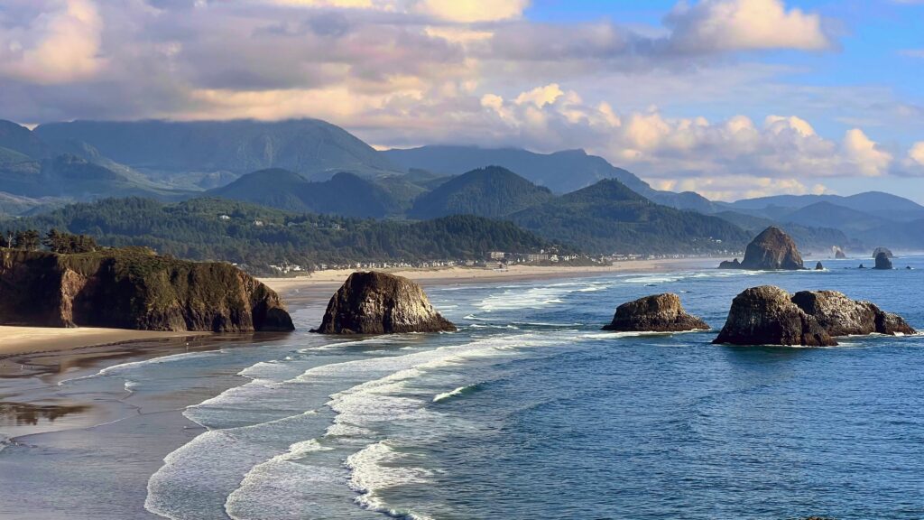 Panoramic view of Cannon Beach, Oregon from Ecola State Park, with sea stacks rising from the surf, a long sandy shoreline, and forested coastal mountains under a partly cloudy sky.