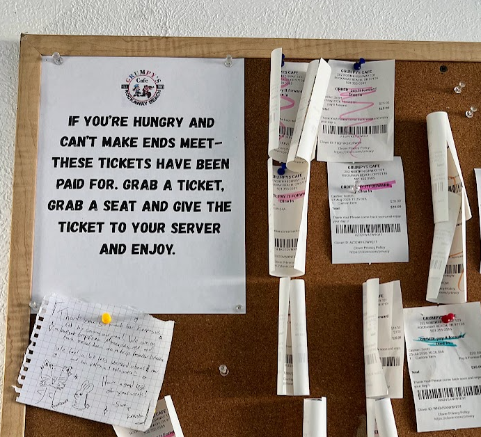 A corkboard at Grumpy's Cafe in Rockaway Beach with a printed sign reading "If you're hungry and can't make ends meet — these tickets have been paid for. Grab a ticket, grab a seat and give the ticket to your server and enjoy," surrounded by pinned prepaid order receipts and a handwritten thank-you note.