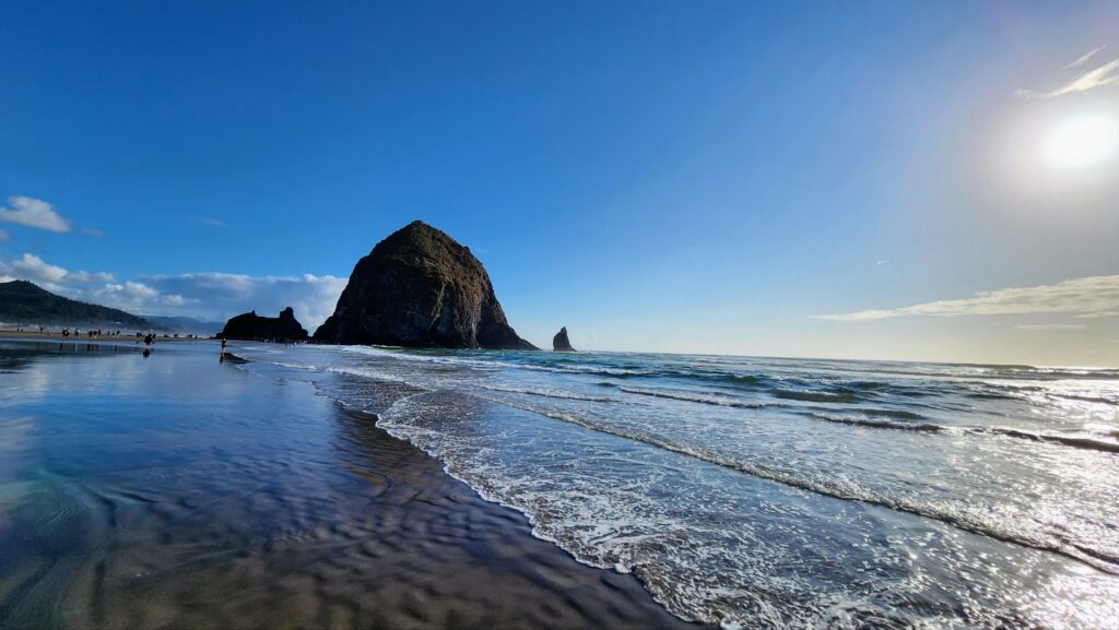 Haystack Rock rising from the shallow surf at Cannon Beach, with wet reflective sand in the foreground, distant people walking along the shore, and a clear blue sky.