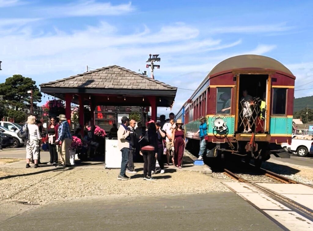 A group of people gathered on a platform beside a vintage passenger train with a maroon, teal, and yellow railcar, next to a covered wooden station shelter decorated with hanging pink flower baskets, under a bright blue sky.