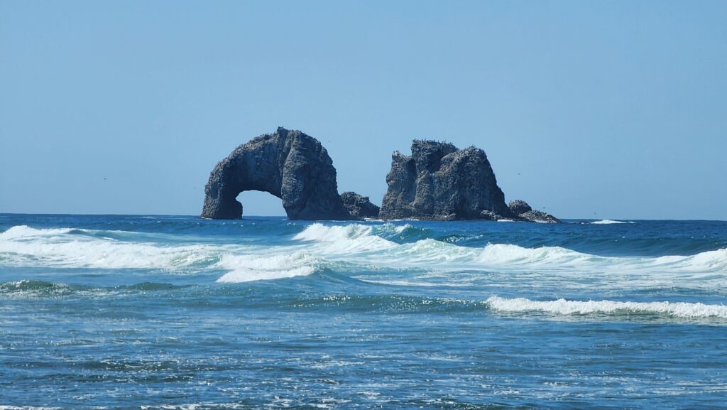 Two large offshore sea stacks under a clear blue sky, the left one pierced by a natural arch, with waves breaking in the foreground.
