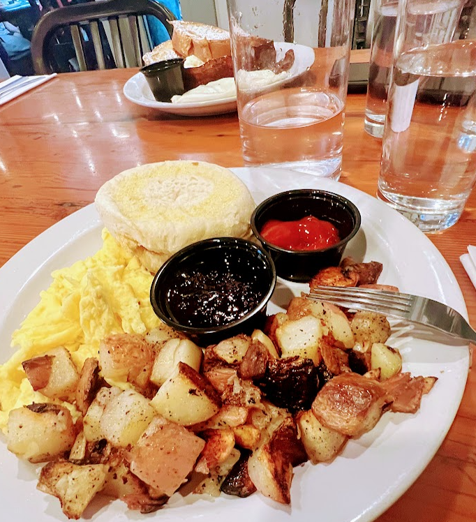 A brunch plate with scrambled eggs, a toasted English muffin, seasoned home-fry potatoes, and small cups of jam and ketchup, on a wooden table with glasses of water and a second plate of food in the background.